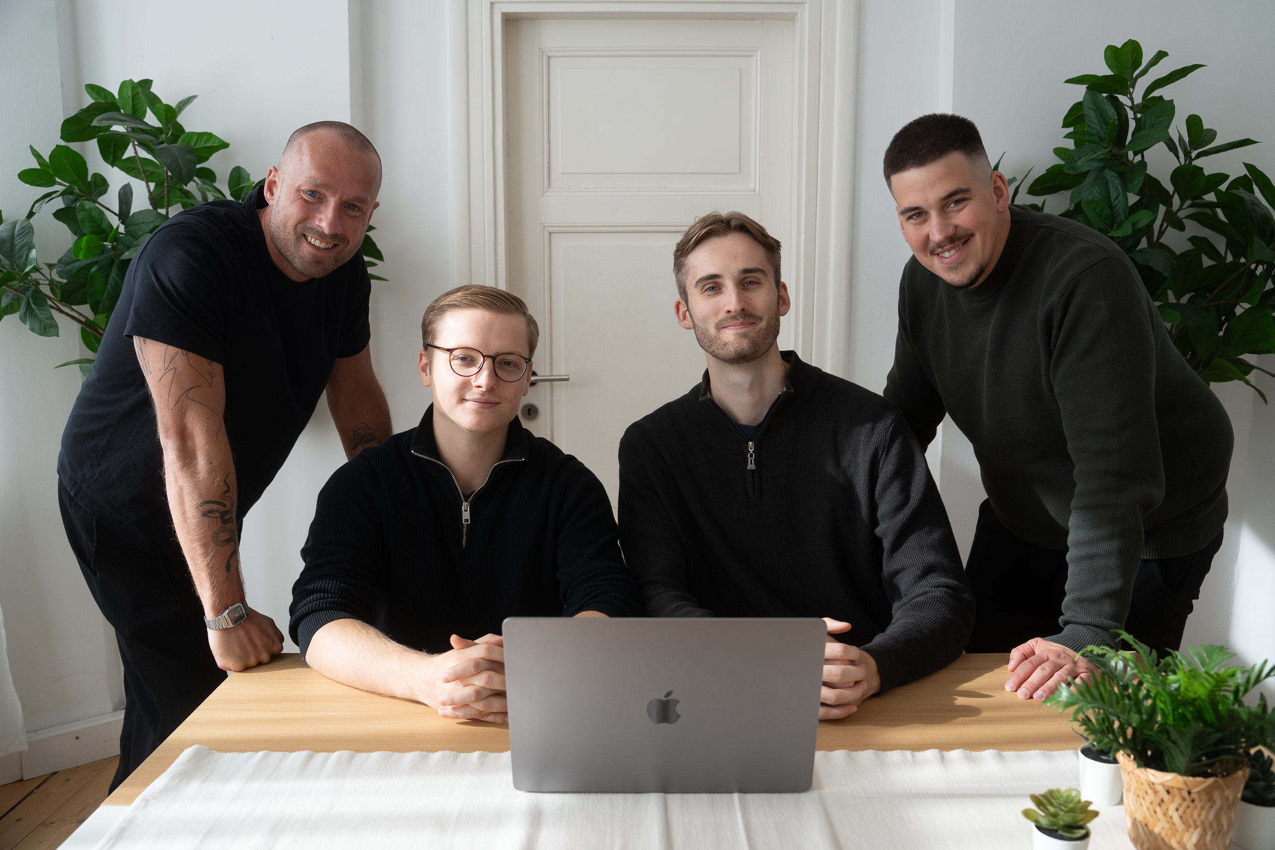 Vier Männer vom Team 360-BPM sitzen und stehen um einen Tisch mit einem geöffneten Laptop, im Hintergrund zwei große grüne Pflanzen.