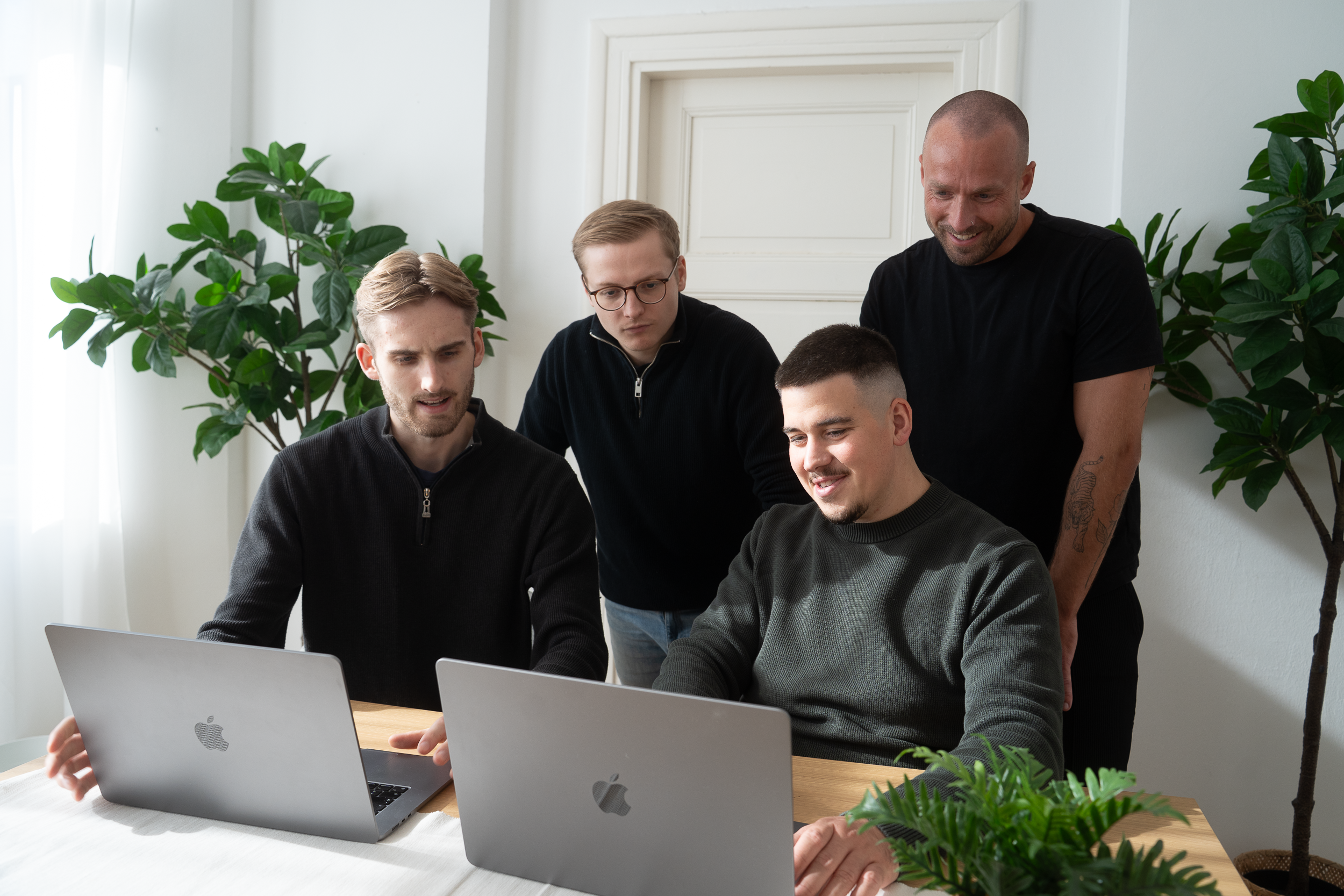 Vier Männer in lässiger Kleidung sitzen und stehen um einen Holztisch mit Laptop und Pflanzen in einem hellen Raum.